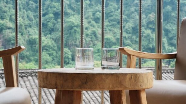 Een houten tafel met twee glazen en uitzicht op een bos door grote ramen.