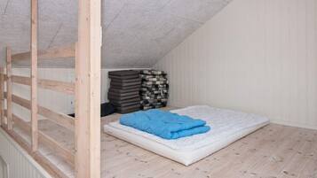 Dormitorio tipo loft con techo inclinado, una escalera de madera, un colchón con una manta azul y una pila de almohadas.