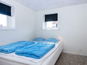 A single bed with a blue blanket and pillows, a window with a view of a house, and a tiled floor.