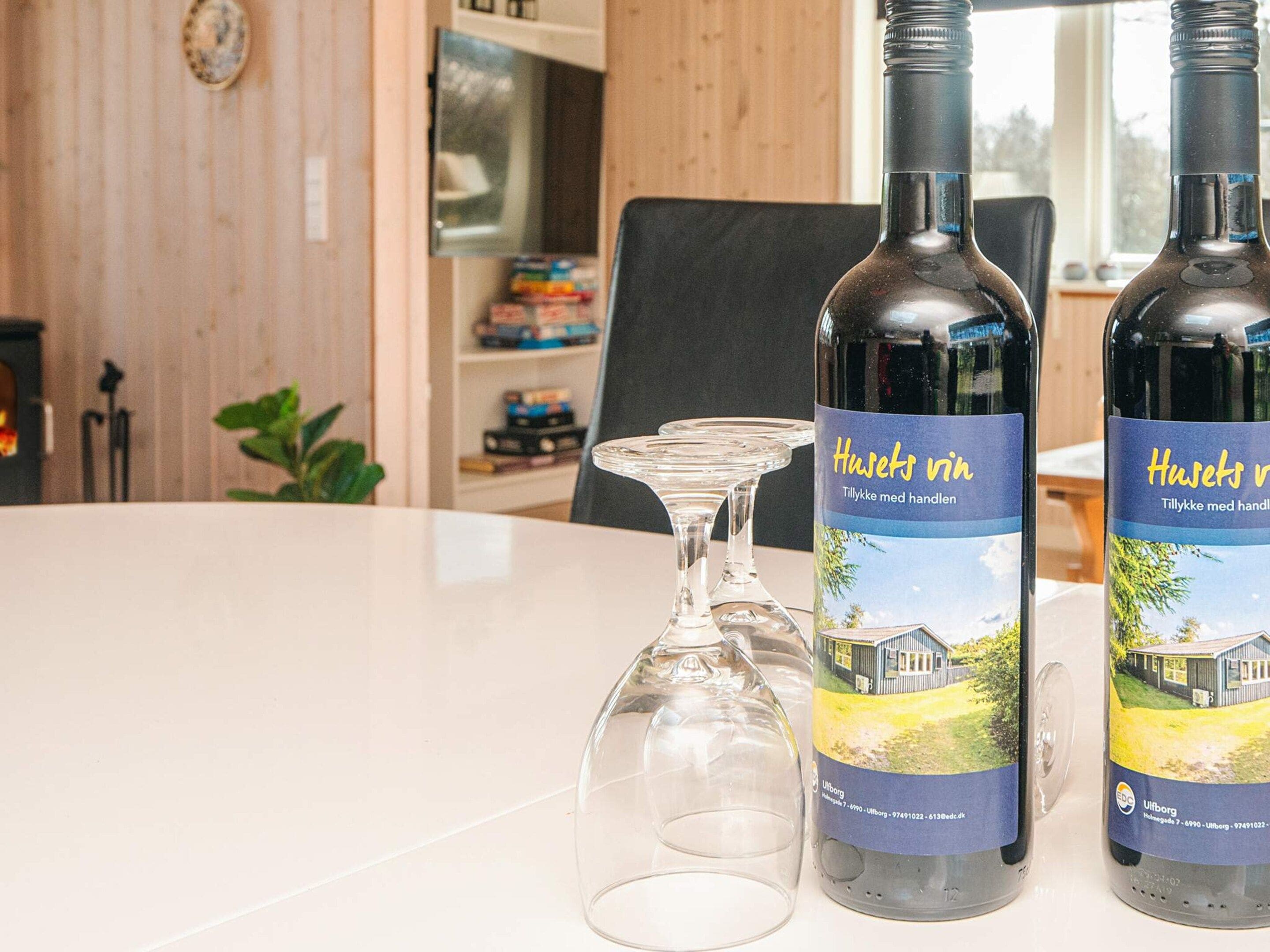Two bottles of Huset vin wine with a label featuring a house and landscape, placed on a table with a clear glass.