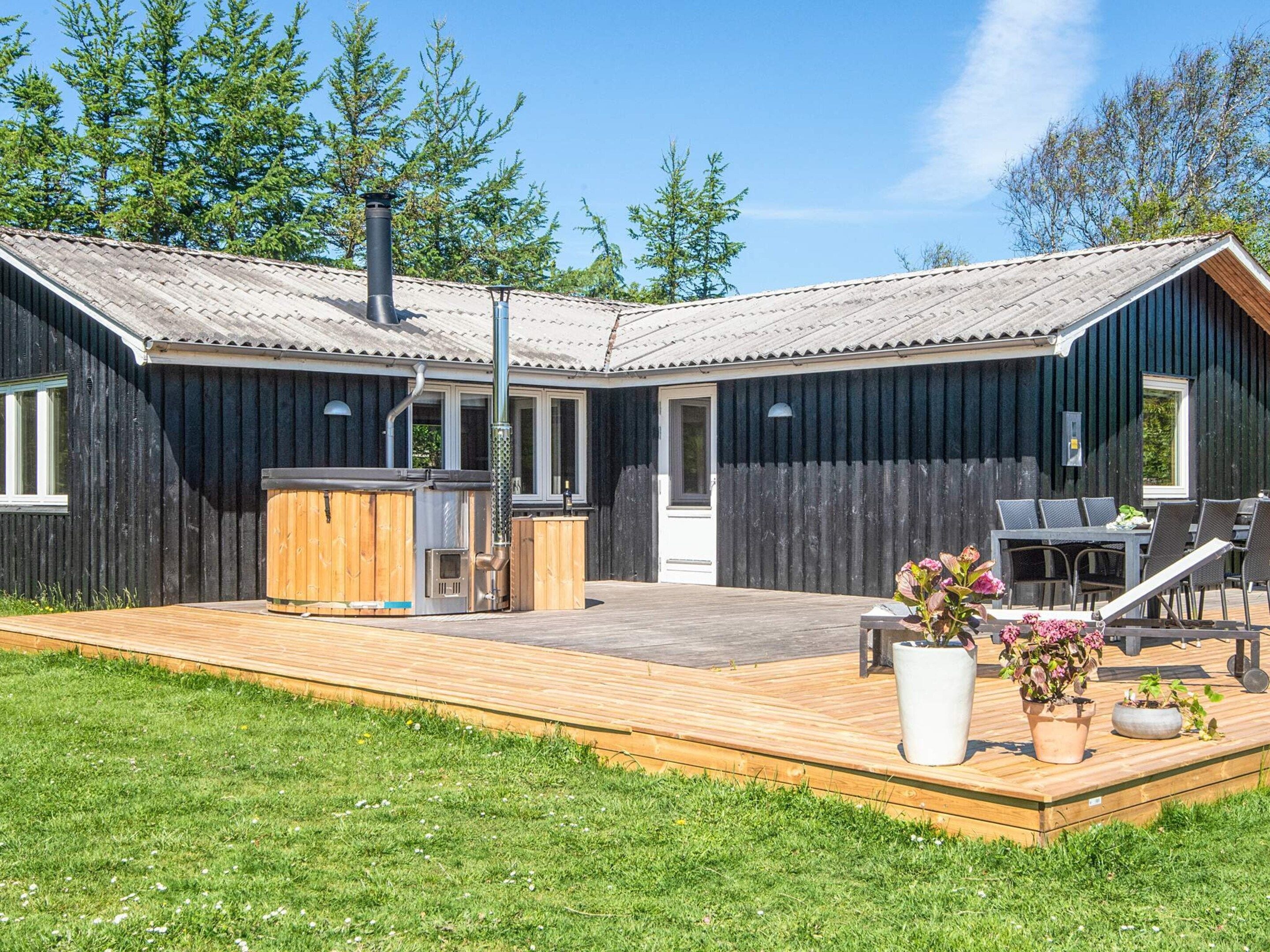Una terraza de madera con jacuzzi, una mesa de patio y sillas, rodeada de césped y árboles.