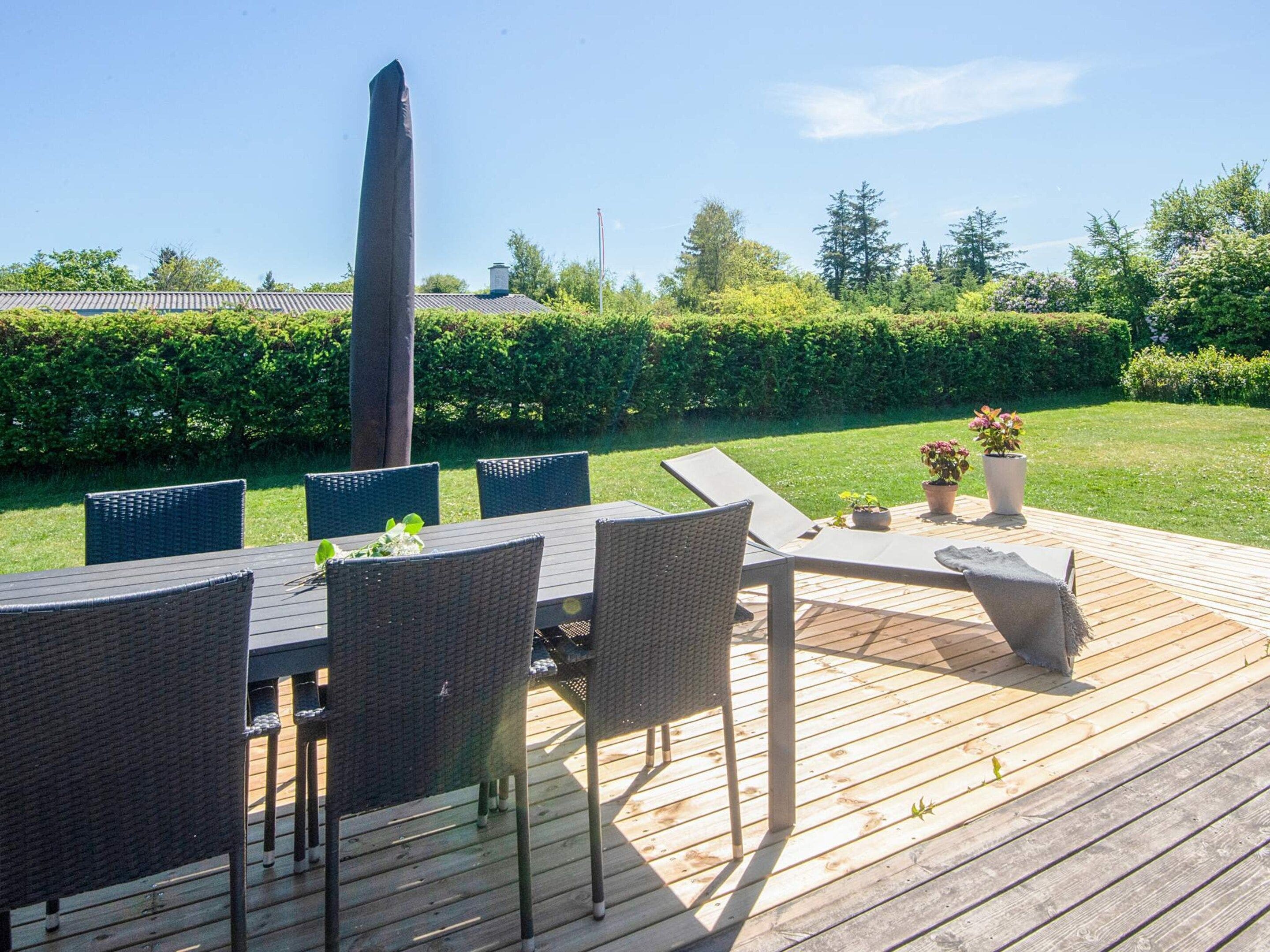An outdoor deck with a dining table and chairs, surrounded by a well-maintained hedge and trees.