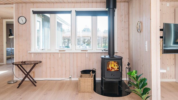 A room with a wood-burning stove, a window with blinds, a clock, and a television on the wall. - 7 Person Holiday Home in Ulfborg (Ulfborg)