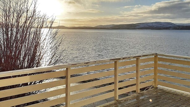 Une terrasse en bois, dotée d’une balustrade, qui offre une vue sur une étendue d’eau calme et sur des montagnes lointaines.