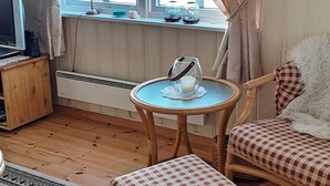 Une pièce chaleureuse avec un plancher en bois, une petite table ronde avec une bougie, une chaise dotée d’un coussin à carreaux et un téléviseur posé sur un meuble en bois.