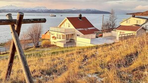 Une maison située sur la côte, avec un toit rouge, une clôture en bois et une vue sur la mer et les montagnes en arrière-plan.