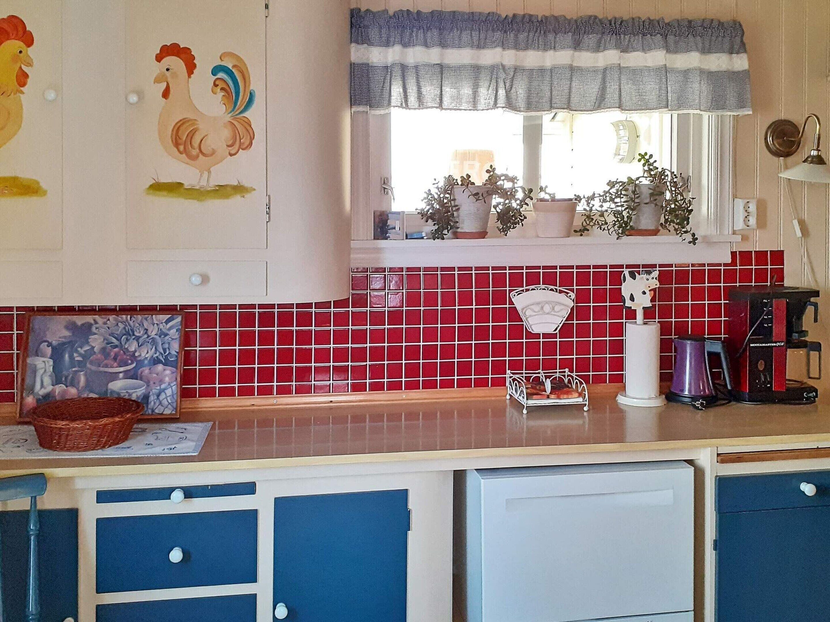 A kitchen with red and white tiles, a window with a checkered valance, and a coffee machine.