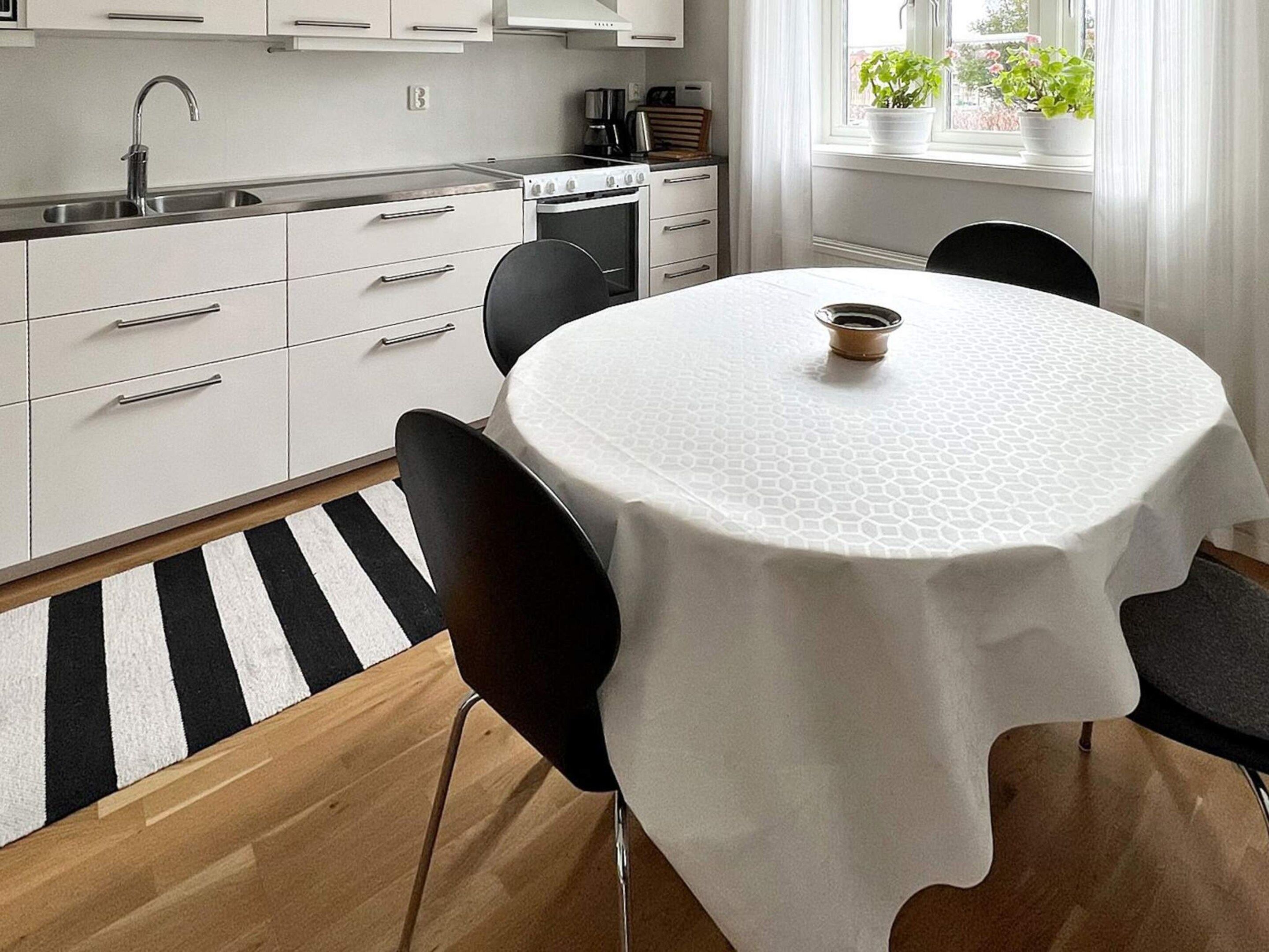 Moderni keittiö, jossa on valkoiset kaapit, ruostumattomasta teräksestä valmistettu pesuallas ja pyöreä pöytä, jonka päällä on valkoinen pöytäliina.