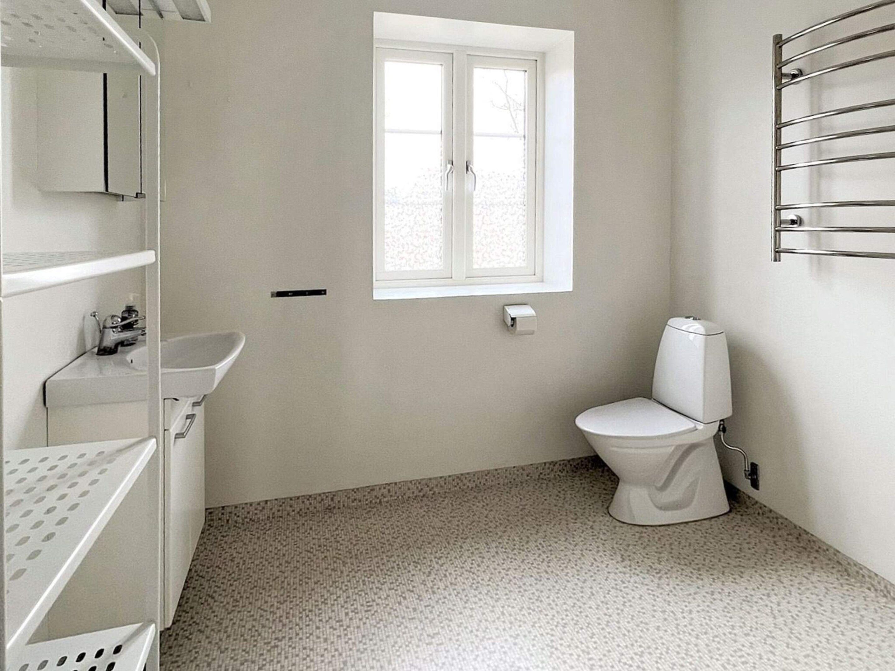 Egy kis, tiszta fürdőszoba, amelyben található egy WC, egy mosdókagyló és egy ablak.