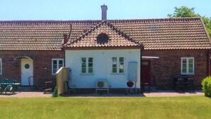 Un bâtiment à deux étages, construit en briques, avec un toit recouvert de tuiles, une porte d’entrée et un petit porche agrémenté de chaises.