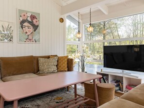 A modern living room with a sofa, a coffee table, and a television. There are bookshelves and a window with a view of trees. - 8 Person Holiday Home in Hals (Hals)