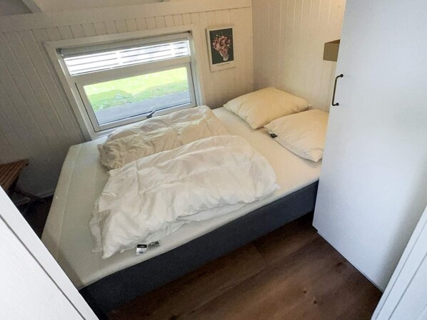 A small, single-bed room with a window, a picture on the wall, and a door to the right.