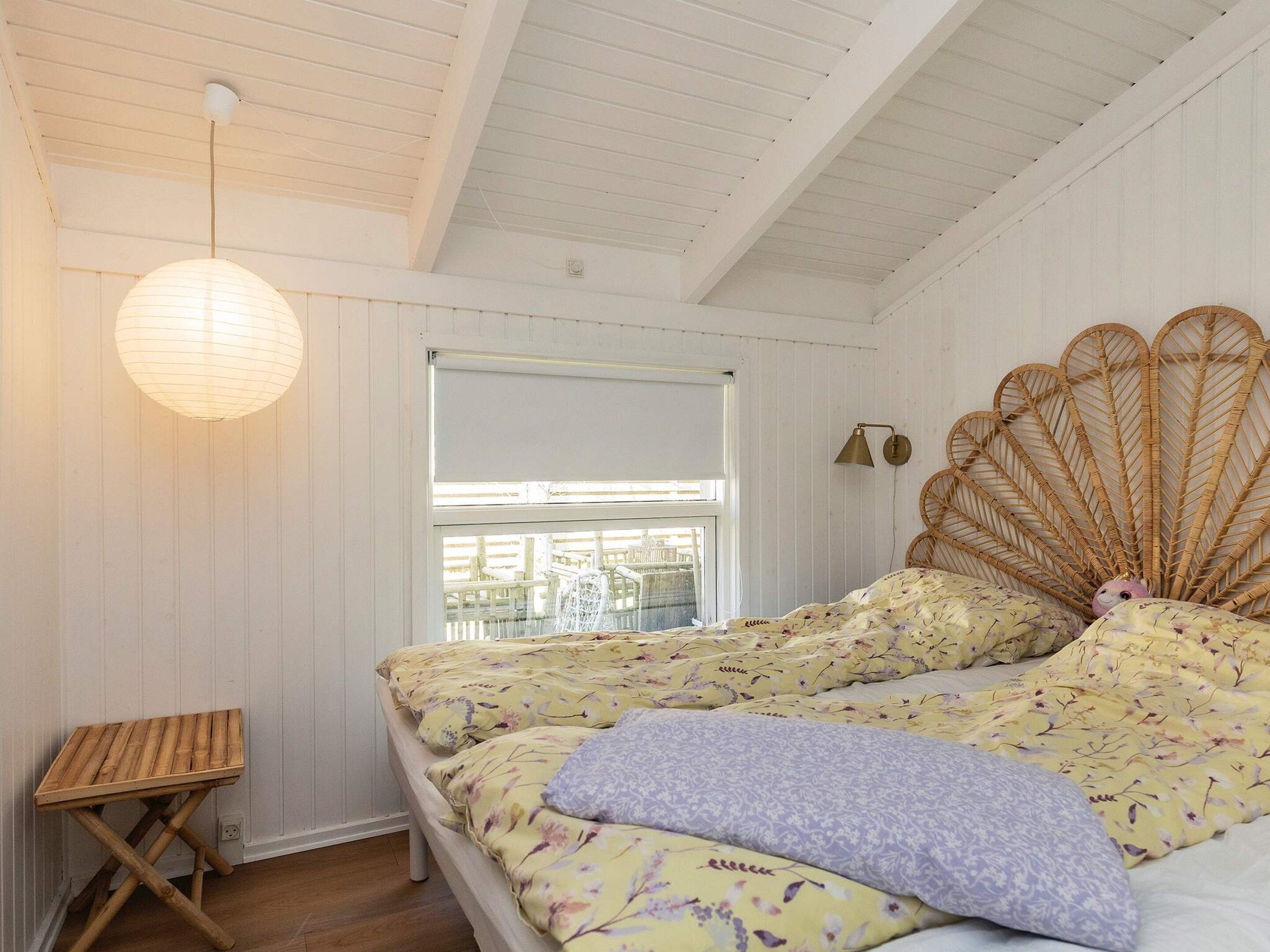 A bedroom with two beds, a wooden stool, a hanging light fixture, and a window with a view of an outdoor deck.