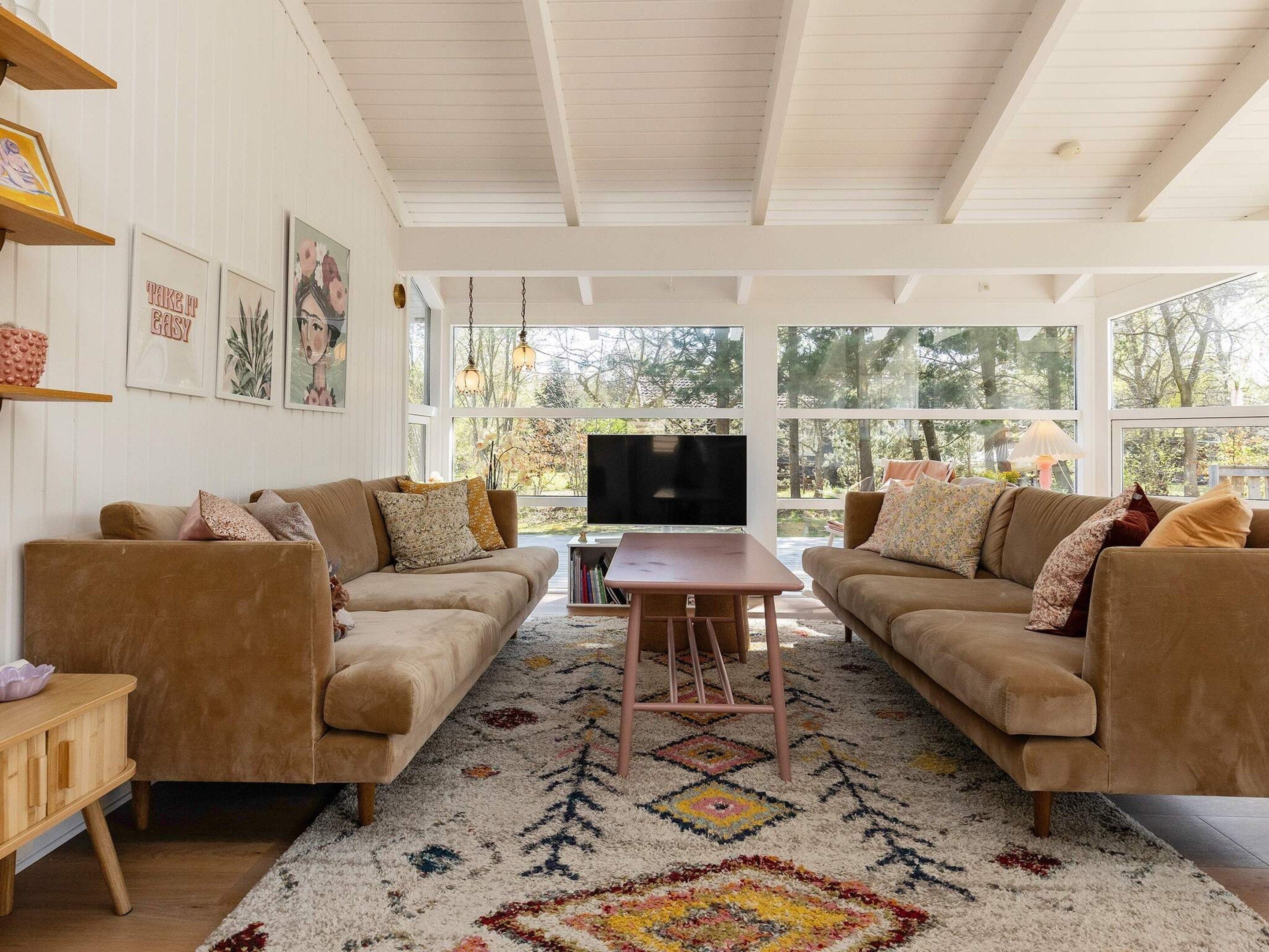 A living room with two sofas, a coffee table, and a TV. There are large windows and a view of trees outside.