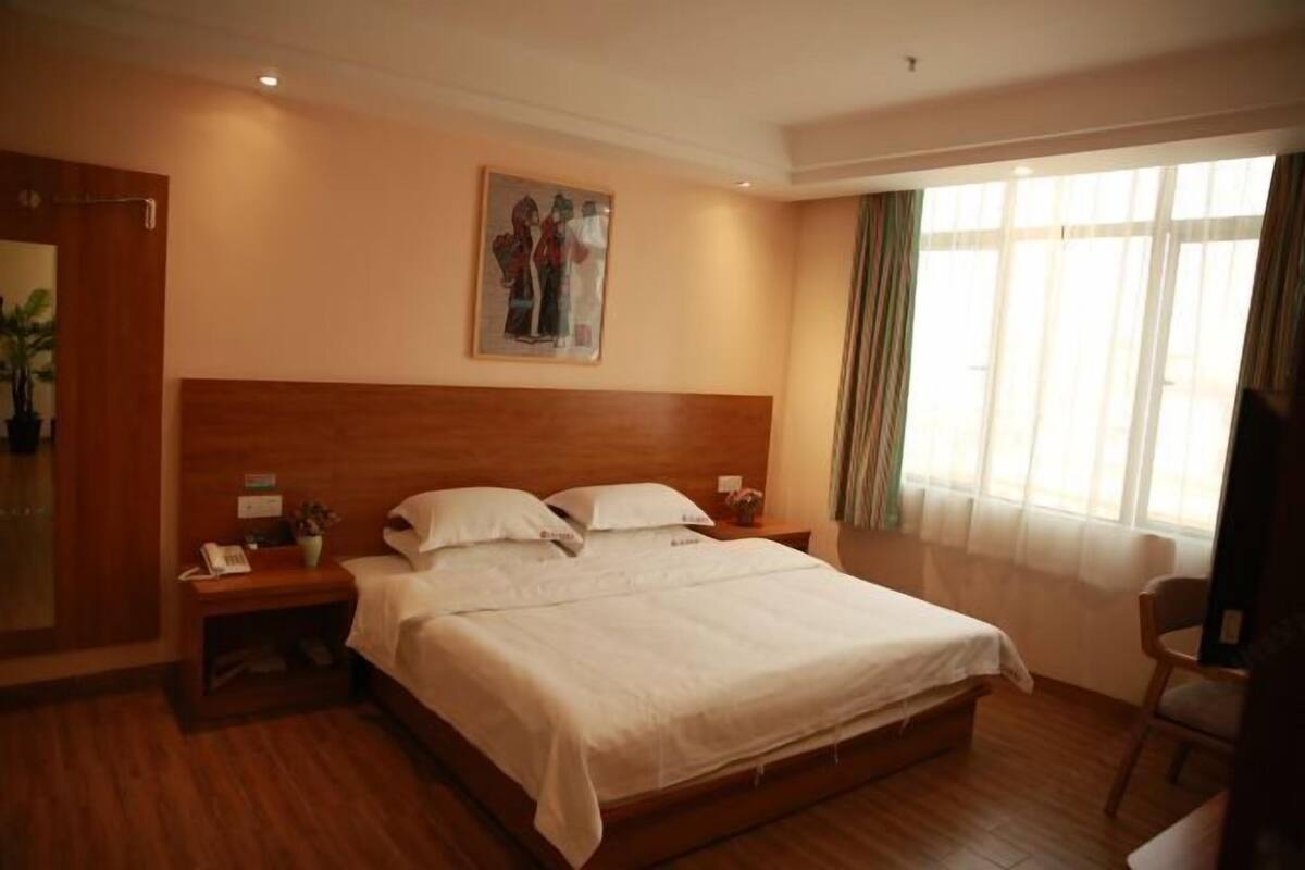 Habitación de hotel con cabecera de madera, cama grande con sábanas blancas, mesita de noche con teléfono y lámpara, un cuadro en la pared y una ventana con cortinas.