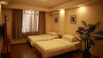 Une chambre d’hôtel avec trois lits, un plancher en bois, une grande fenêtre avec des rideaux et une plante en pot.