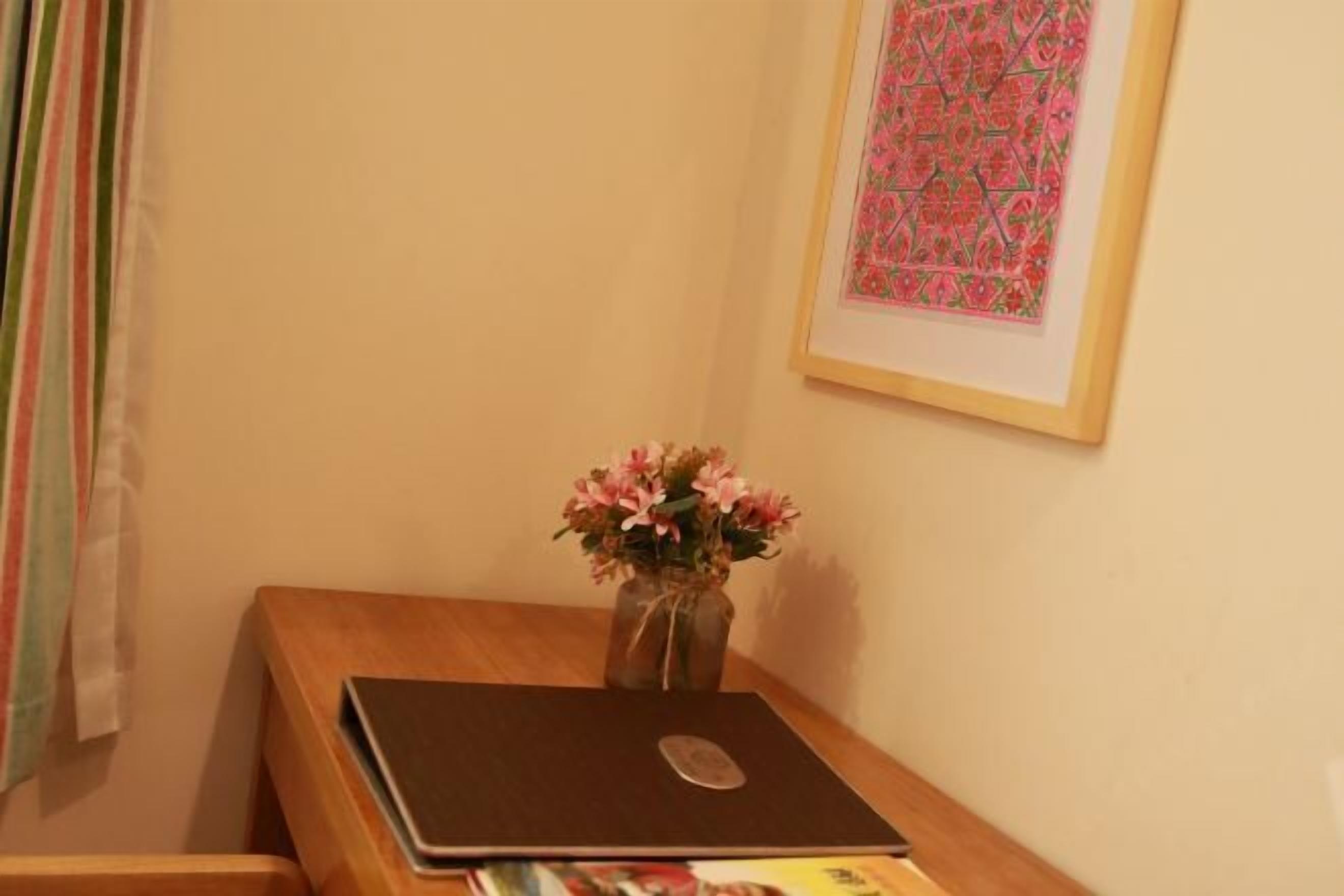 Um computador portátil sobre uma escrivaninha de madeira, com um vaso de flores, um quadro na parede e uma janela com cortinas.
