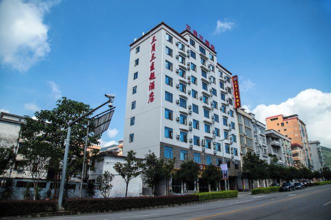 Un hotel de varios pisos con un llamativo letrero rojo en la fachada, un cielo azul despejado y una calle con coches estacionados y vegetación.
