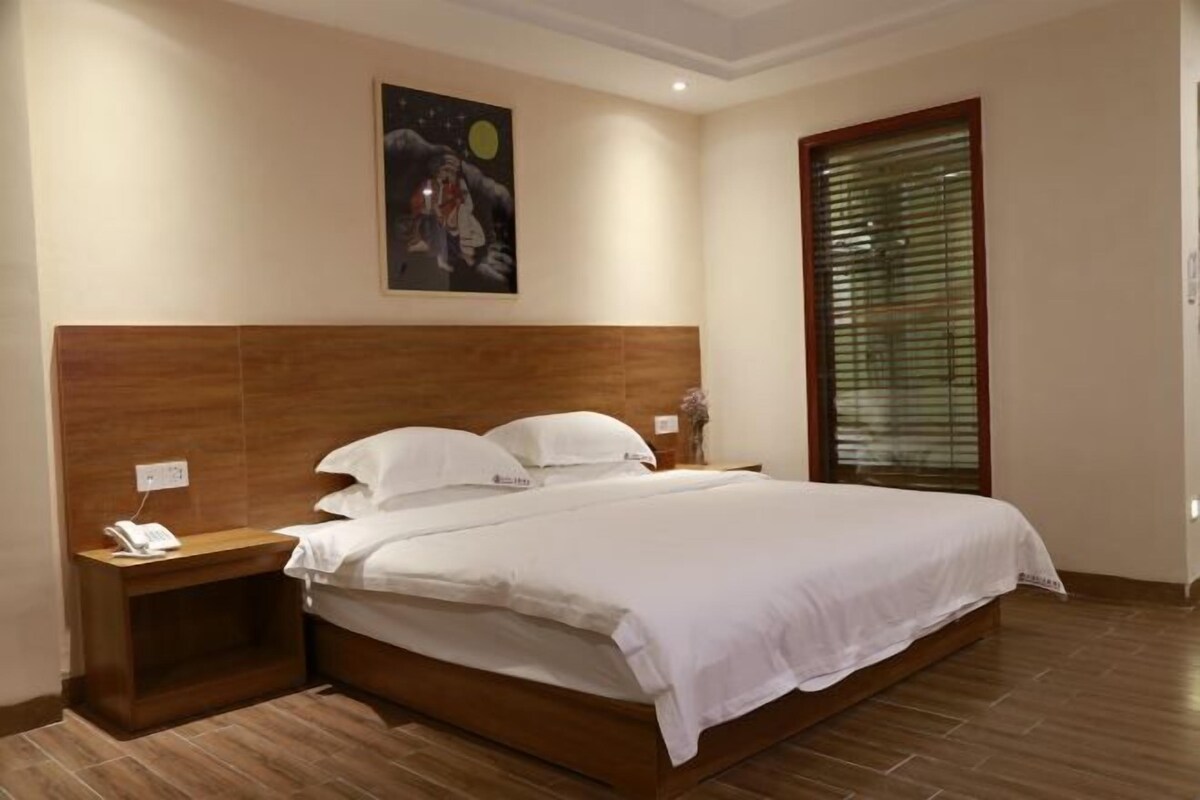 Un dormitorio con cabecera de madera, colcha blanca, mesita de noche con un teléfono y ventana con persianas.