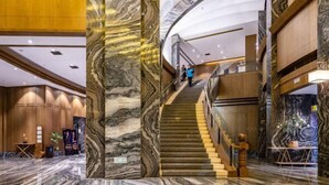 Um amplo saguão de hotel com paredes de mármore, uma escadaria central e um balcão de recepção.
