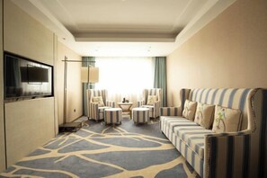 Une chambre d’hôtel avec un canapé, des fauteuils, une télévision et une grande fenêtre avec des rideaux.