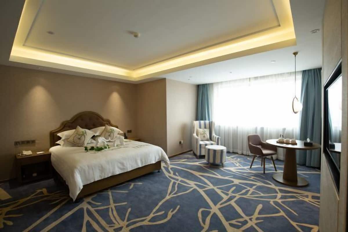 A hotel room with a large bed, a round table, two chairs, a small round table, and a large window with curtains.