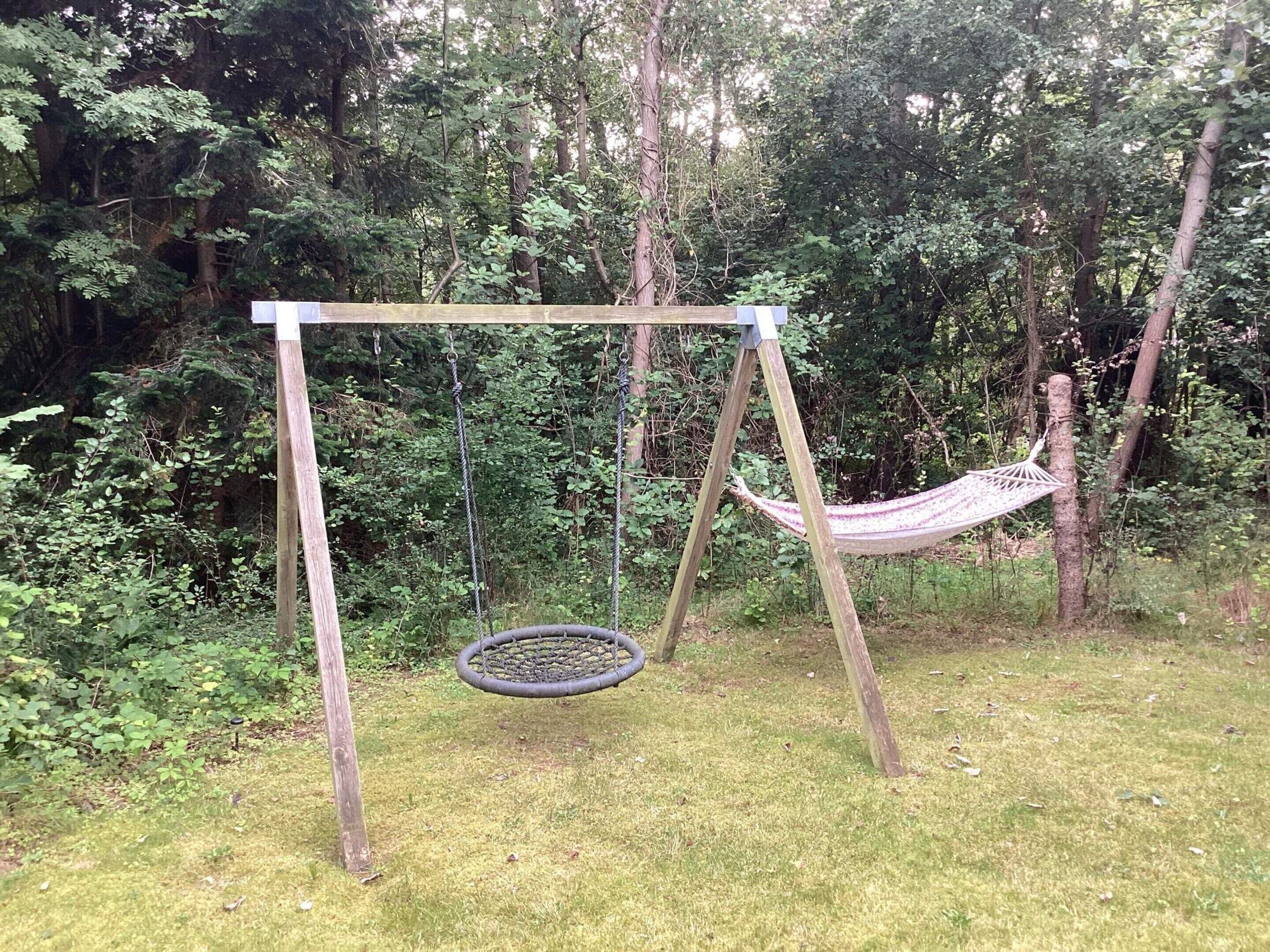 A wooden swing set with a hammock in a wooded area.