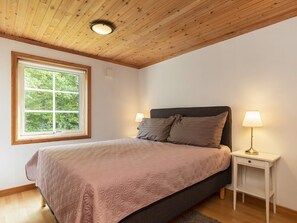 A bedroom with a bed, a nightstand with a lamp, a window with a view of trees, and a wooden ceiling.