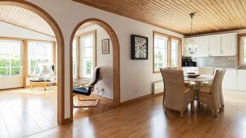 A spacious room with wooden flooring, a dining area with wicker chairs, and large windows allowing natural light.