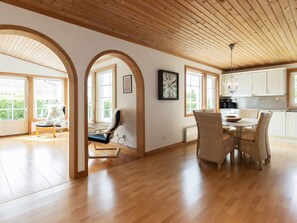 A spacious room with wooden flooring, a dining area with wicker chairs, and large windows allowing natural light.