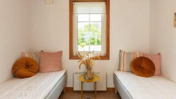 Quarto pequeno com duas camas de solteiro, janela com vista para área verde, radiador e pequena mesa com vaso de plantas secas.