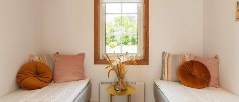 A small room with two single beds, a window with a view of greenery, a radiator, and a small table with a vase of dried plants.