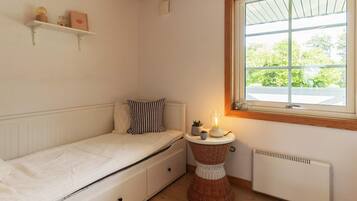 A small, cozy room with a single bed, a bedside table with a lamp, a radiator, and a window with a view of greenery.