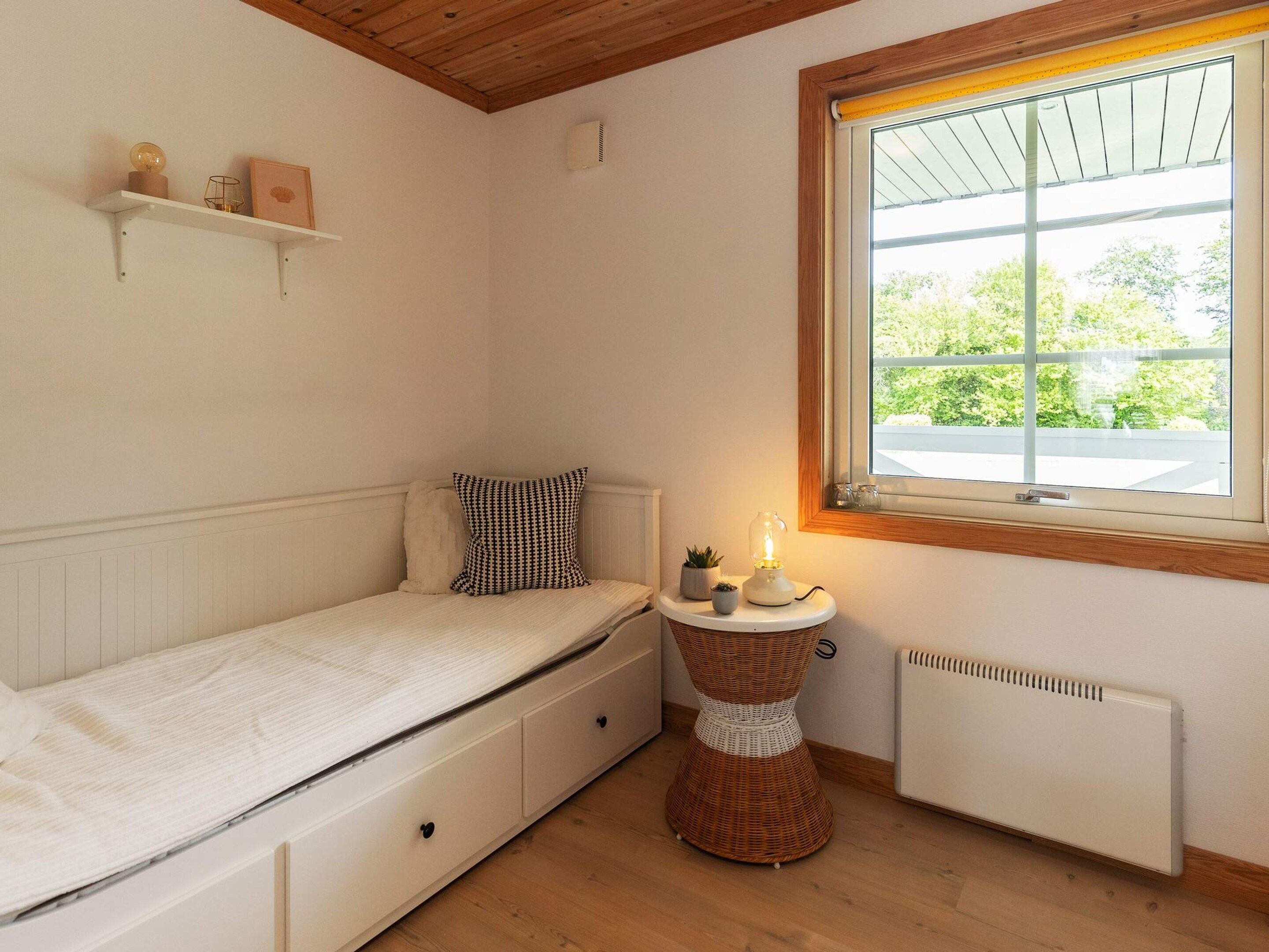 A small bedroom with a single bed, a bedside table with a lamp, a radiator, and a window with a view of greenery.