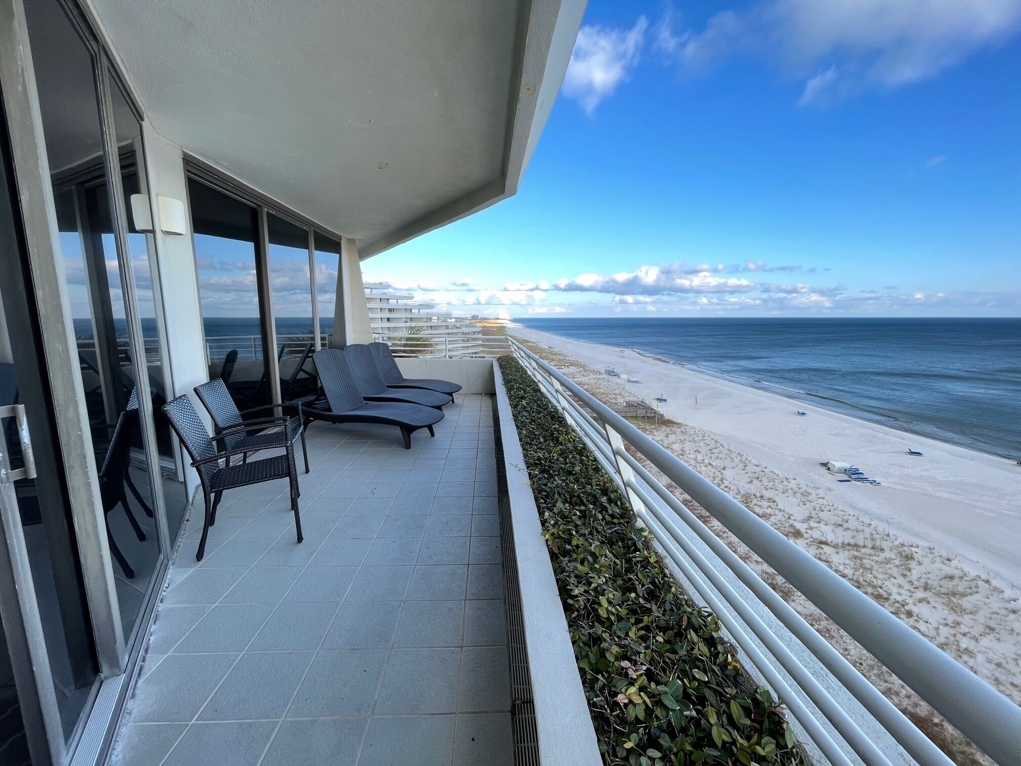 A balcony with two lounge chairs and a table, overlooking a beach with a railing and a clear view of the ocean.