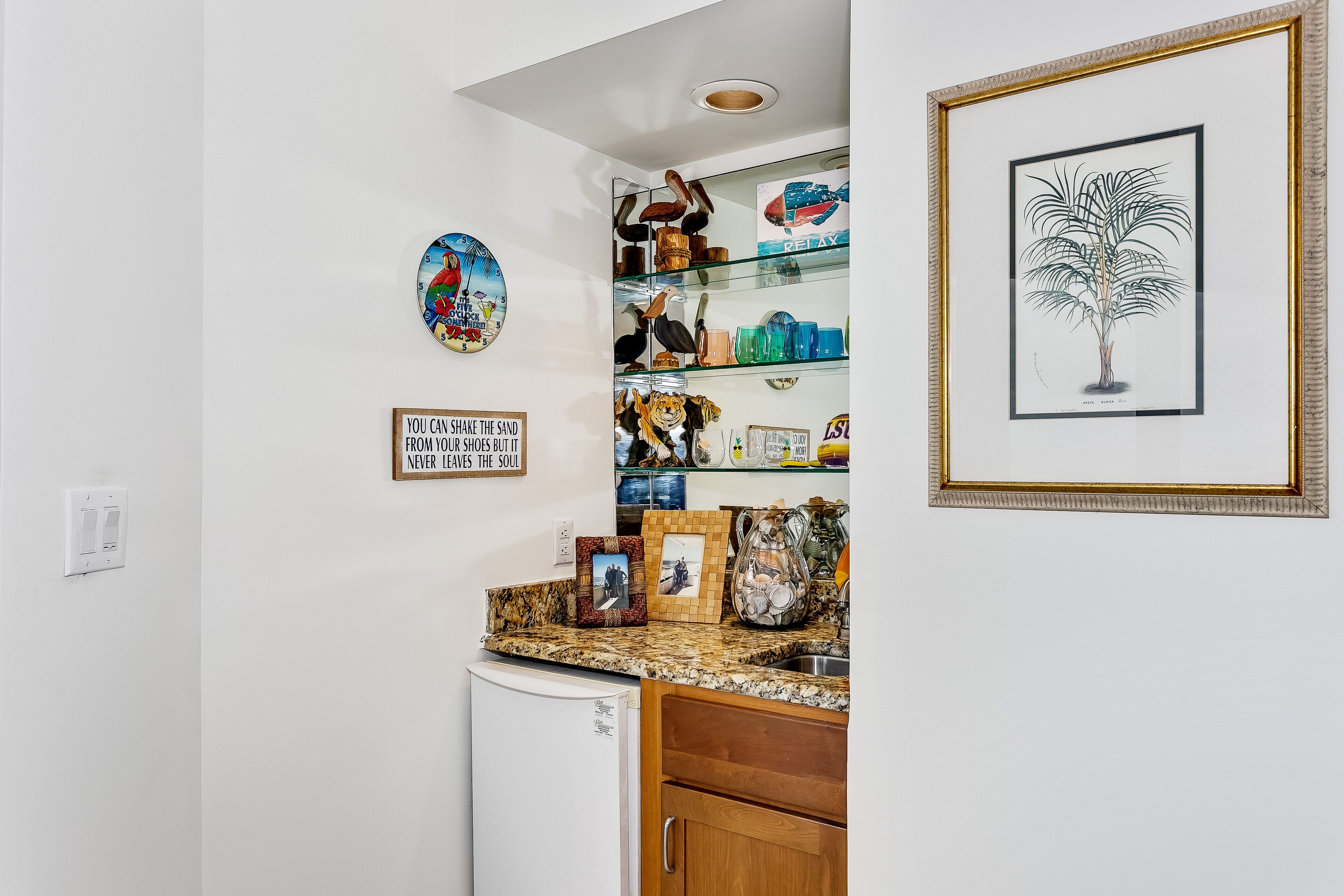 A kitchen with a white refrigerator, wooden cabinets, a granite countertop, and various decorative items including a framed plant, a clock, and a sign.