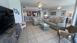 A spacious living room with a TV, sofa, coffee table, and wicker chairs. A kitchen area is visible in the background.