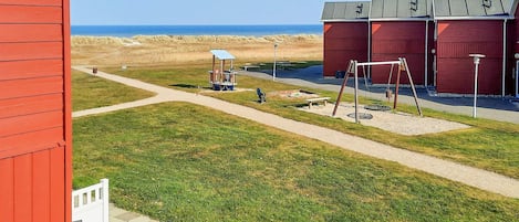 Un edificio rojo con una valla blanca, un parque infantil y vista a la playa.