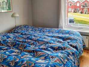 A room with a bed covered in a patterned blanket, a window with a view of a red building, and a wall-mounted lamp. - 6 Person Holiday Home in Hadsund (Hadsund)