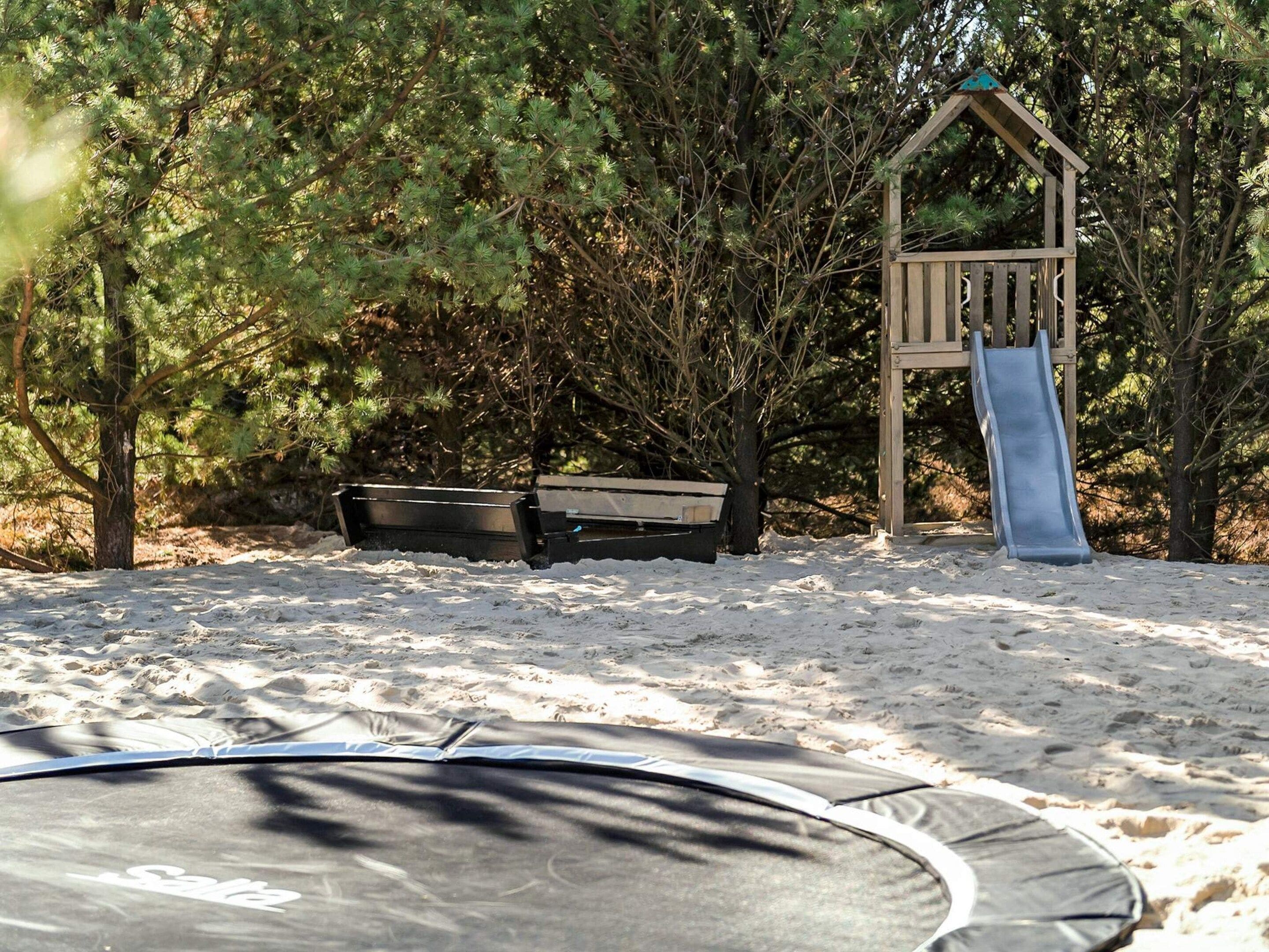 A playground with a slide, a swing set, and a trampoline in a wooded area.