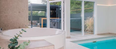 A modern indoor pool area with a large jacuzzi, glass doors leading to an outdoor deck, and a view of the surrounding landscape.