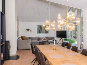 A modern living room with a grey sofa, a wooden dining table with chairs, and a large window with a view of trees.