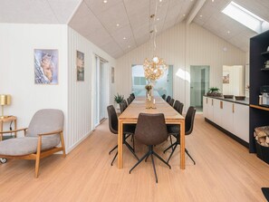 A modern dining room with a long wooden table, black chairs, a chandelier, and a kitchen area with cabinets and a countertop.