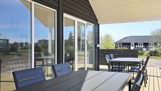 An outdoor patio with a table and chairs, large sliding glass doors, and a view of a house in the background. - 8 Person Holiday Home in Vaeggerlose (Vaeggerlose)