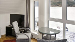 A room with a large window overlooking a body of water, wicker chairs, a small glass table, a TV on a stand, and a red patterned rug. - 6 Person Holiday Home in Gursken-by Traum (Sande)