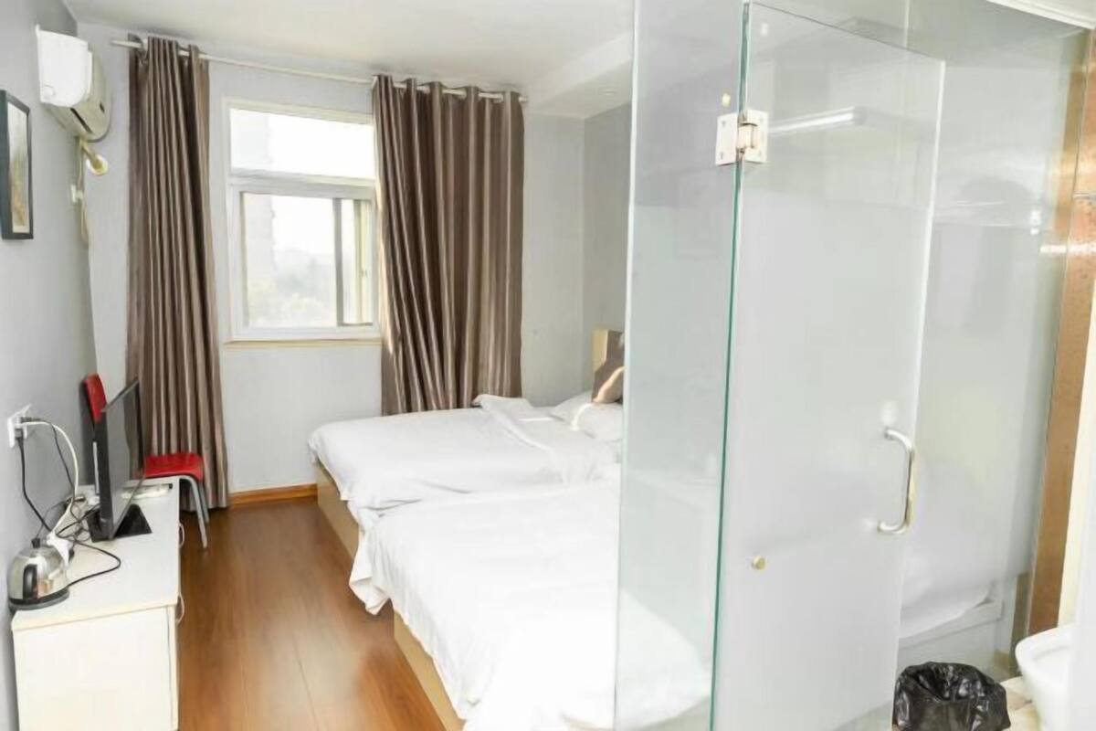 A hotel room with a bed, a desk with a computer, a window with curtains, and a glass shower enclosure.