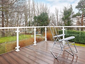 A metal folding chair on a wooden deck with a white railing, overlooking a grassy area with trees and a hedge.