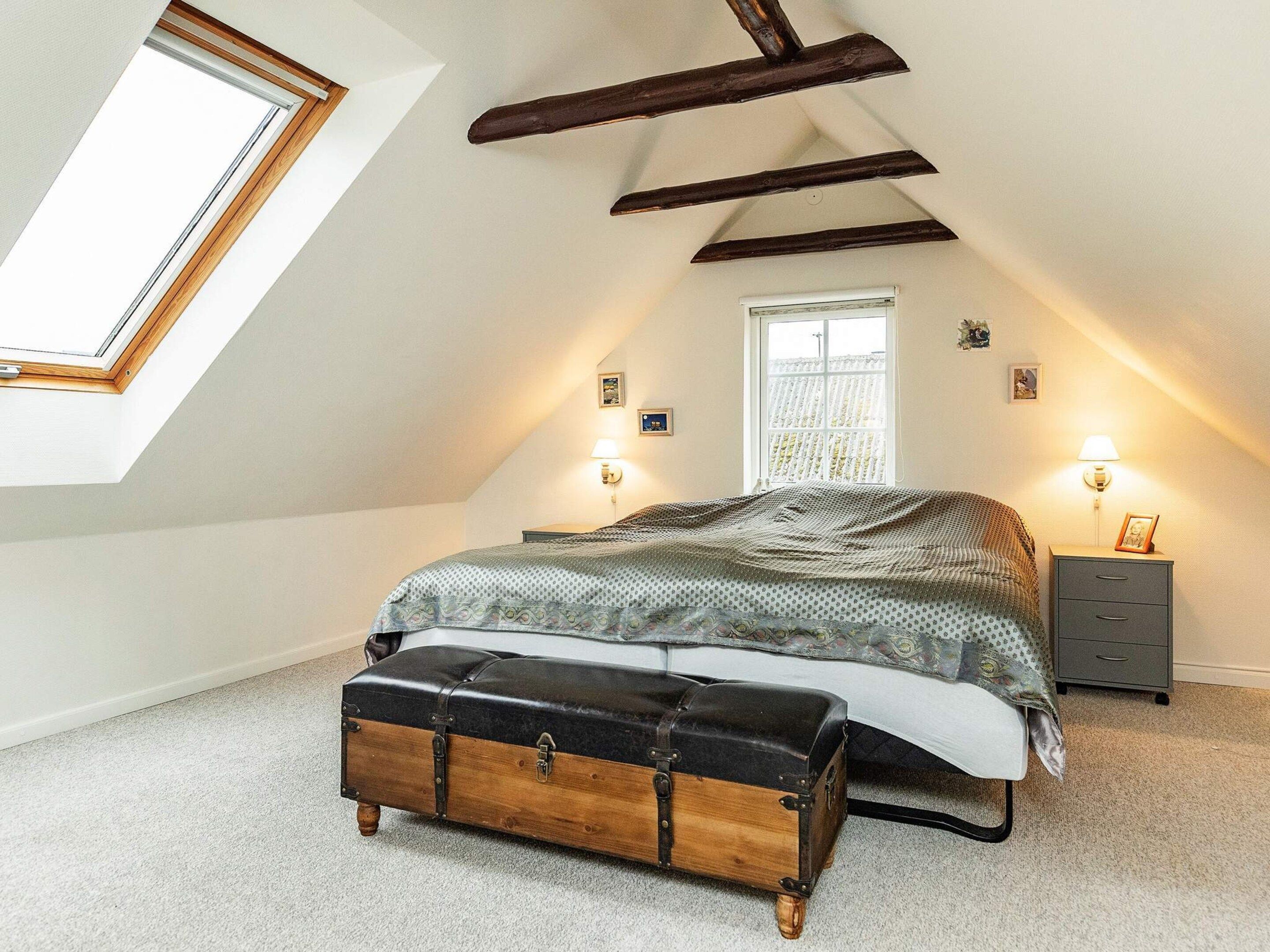 A bedroom with a sloped ceiling, a skylight, a bed with a patterned bedspread, a bench, a nightstand with a lamp, and a window with curtains.