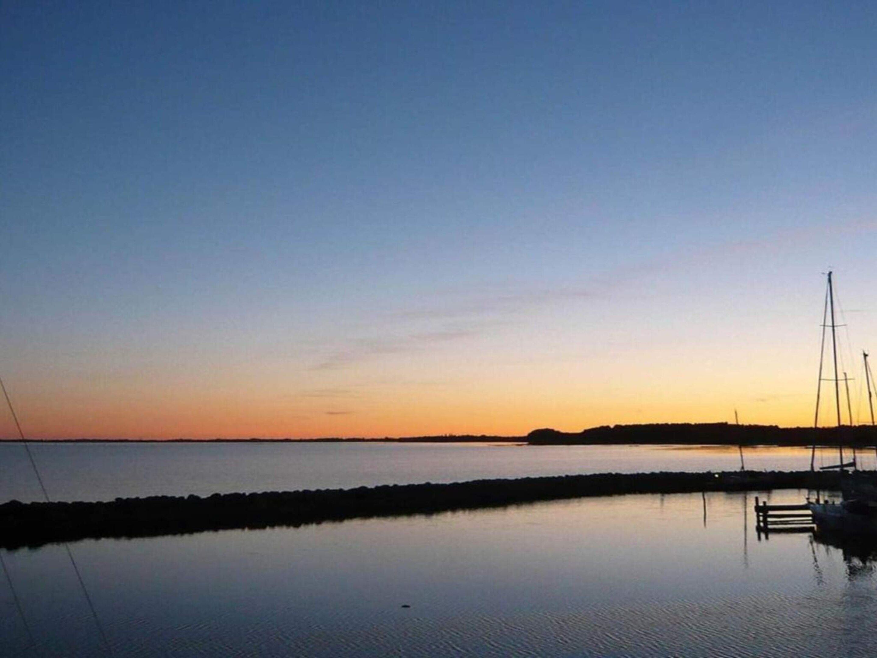 A calm body of water reflecting the sky during sunset, with a dock and sailboats in the distance.