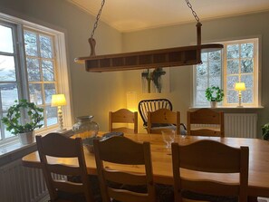 Comedor con mesa y sillas de madera, lámpara colgante y vistas a árboles a través de las ventanas.